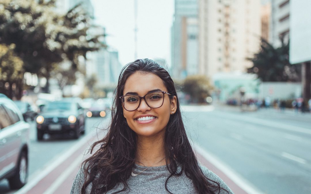 A woman wearing large black rimmed glasses standing in a busy city street smiles broadly.