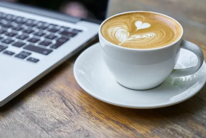 Mug of latte art on a white saucer, placed next to a silver laptop keyboard.