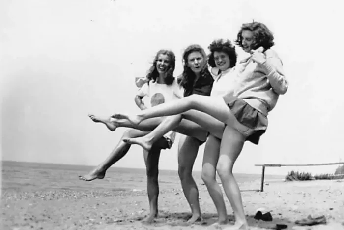 A black and white photo of four young women posing on a beach with a calm ocean in the background.