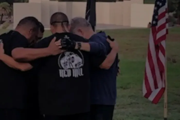 A group of people are seen from behind, standing close together in an emotional embrace on a grassy area.