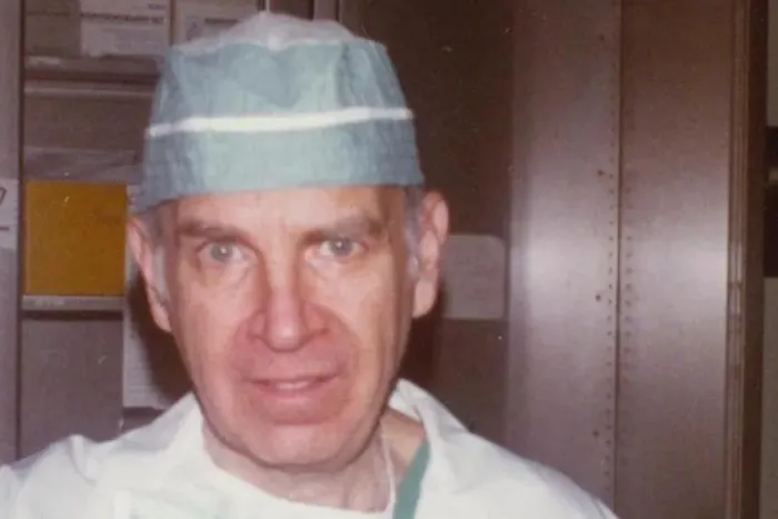 A color portrait of an older man, likely a doctor or surgeon, wearing a light green surgical cap and a white medical coat.